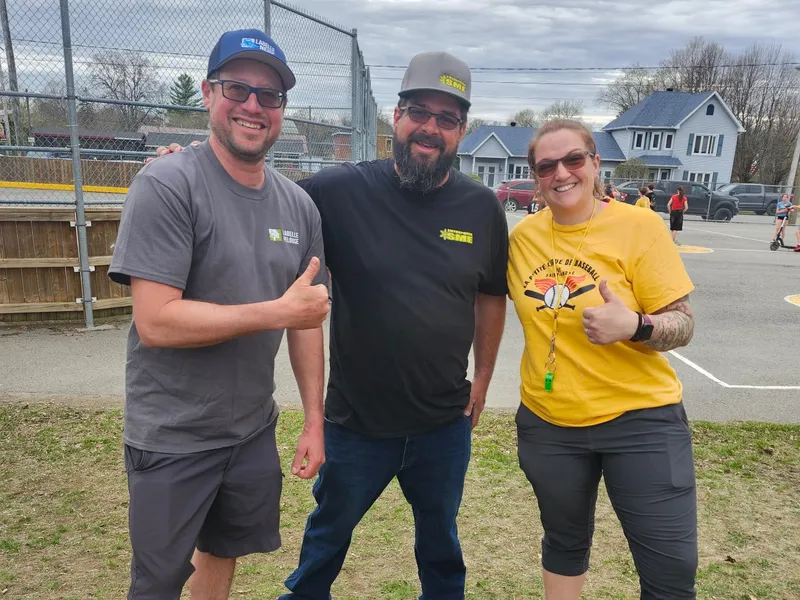Simon Constantin en chandail Entreprise SME avec deux personnes devant le terrain de baseball, pouces levés